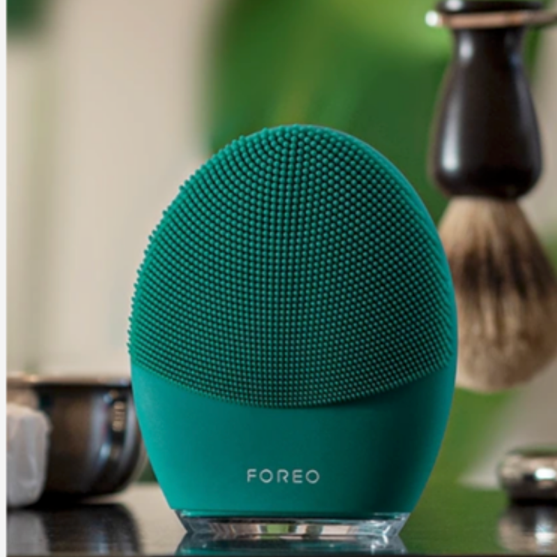 The front side of the FOREO LUNA 4 MEN smart facial brush in Evergreen, displayed on a bathroom counter alongside a classic shaving brush.