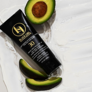 Flat lay photograph of a black tube of Black Girl Sunscreen Moisturizing Sunscreen Lotion SPF 30 resting on a textured background of swirled white lotion, artistically surrounded by fresh avocado slices and a halved avocado with its pit intact.