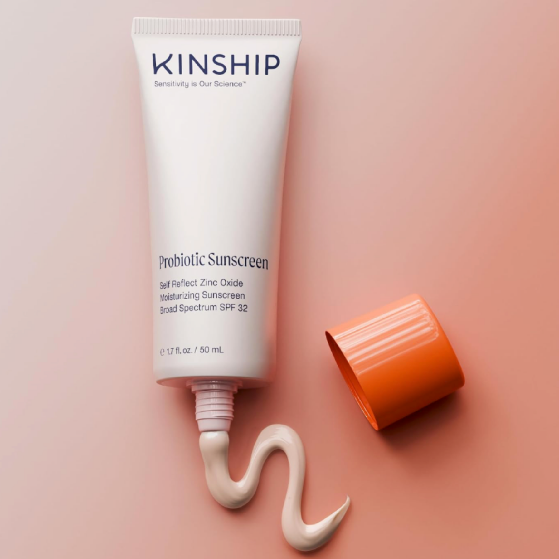 Close-up product shot of a white Kinship sunscreen tube with its orange cap removed, squeezing a thick ribbon of light-pink creamy sunscreen texture onto a soft peach background.