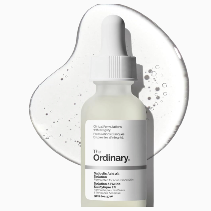 Product shot of The Ordinary Salicylic Acid 2% Solution in a frosted glass dropper bottle set against a large, clear liquid serum puddle with tiny air bubbles. The bottle label describes the formula as a clinical treatment designed for acne-prone skin.