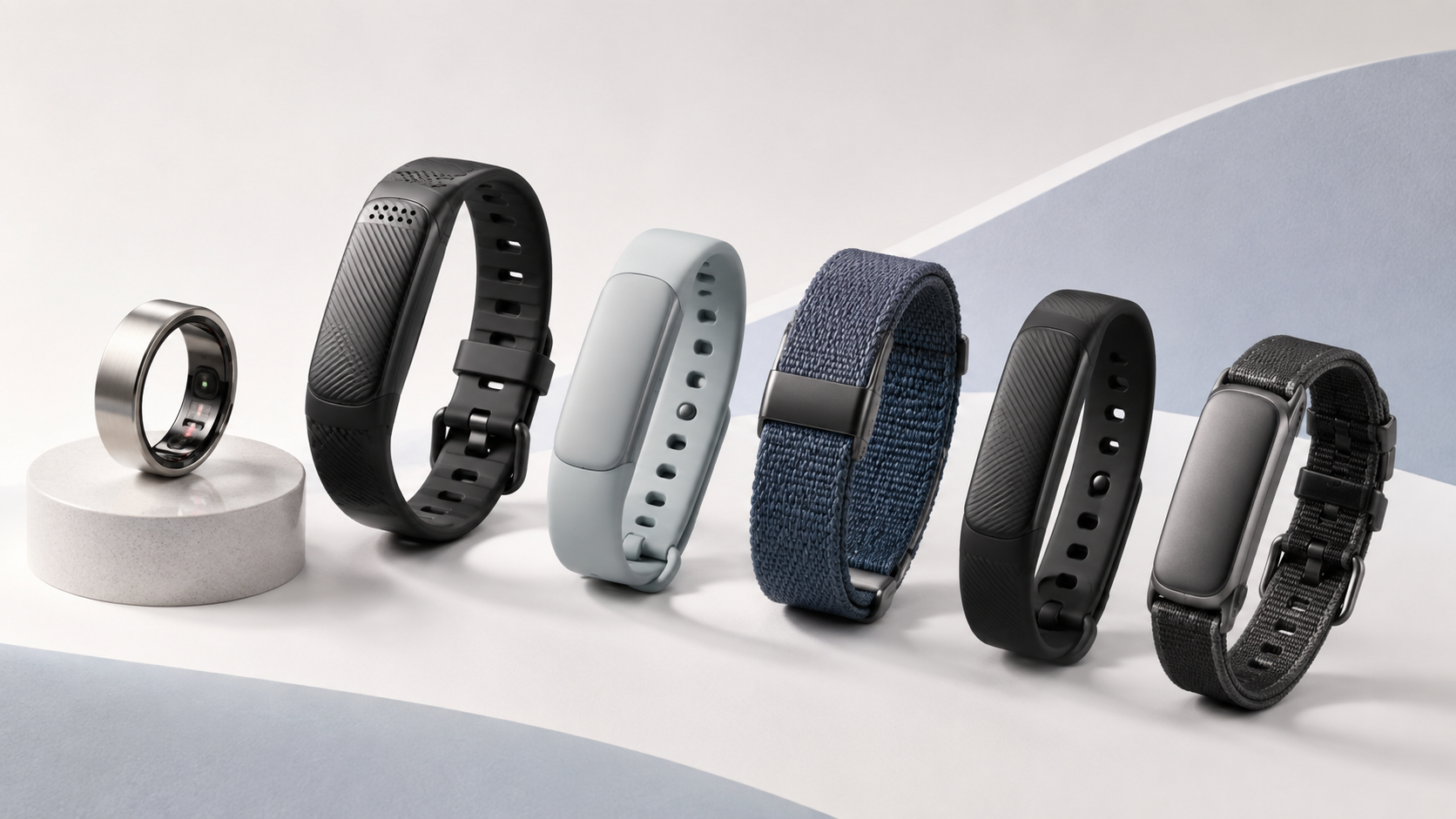 Editorial display of six screen-free health and fitness wearables on a curved white platform with a soft gray-blue backdrop. The collection includes a silver smart ring with visible inner sensors and five screen-free tracking bands in black, pale blue, navy woven fabric and charcoal styles.